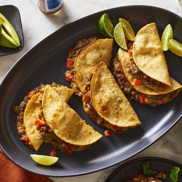 Fried tacos filled with meat and peppers on a dark platter, served with avocado, lime, and beer.