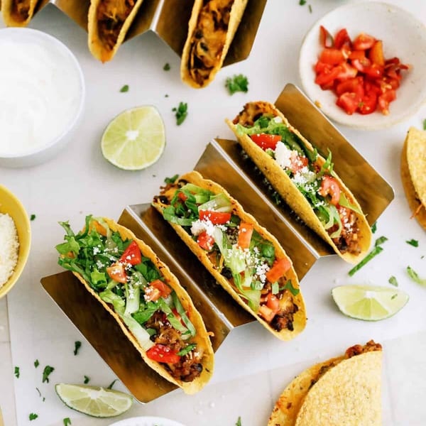 Crispy hard-shell tacos in metal holders, some filled with fresh lettuce, tomatoes, and cheese, alongside lime.